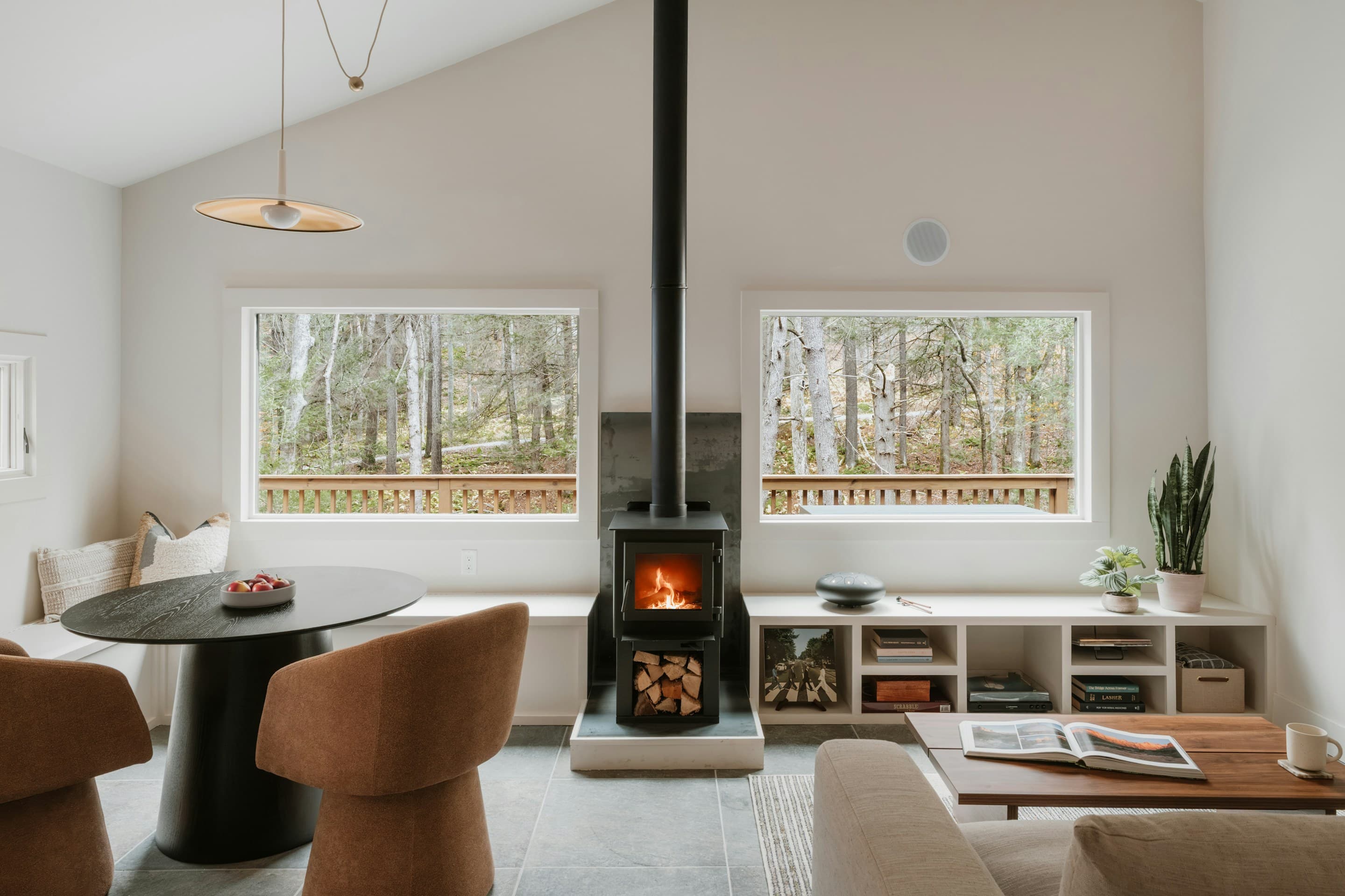 Minimalist cabin great room with vaulted ceiling, black wood-burning stove flanked by twin windows, dining nook, and rust-colored upholstered chairs